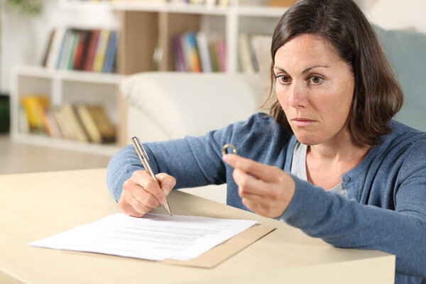 Pensive wife signing divorce papers looking away holding wedding ring sitting in the livingroom at home