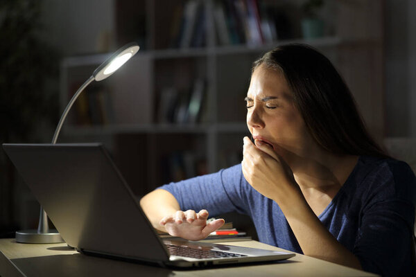 Tired woman with laptop yawning sitting late with a desk lamp on the table at night at home