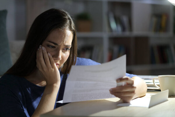 Worried woman reading letter sitting alone in the living room at night at home