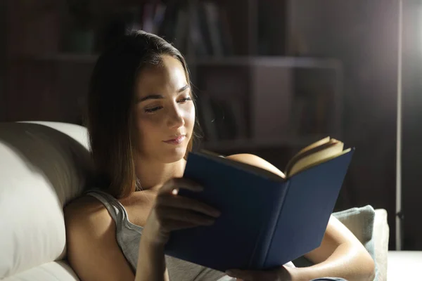 Relaxed woman reading a paper book sitting on a couch at home at night