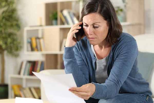 Serious adult woman calling on smart phone checking letter sitting on the sofa at home