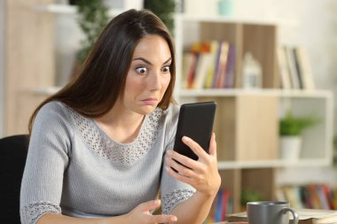 Kafası karışık bir kadın akıllı telefonu kontrol ediyor evdeki oturma odasında masanın üstünde oturuyor.
