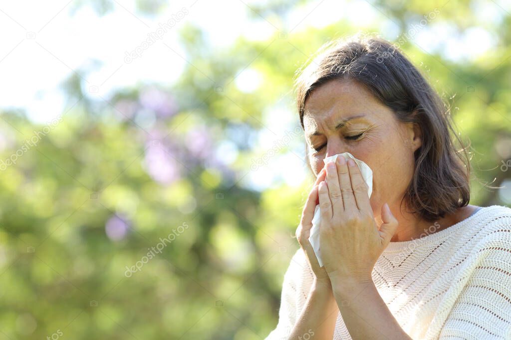 Sick adult woman blowing nose with tissue standing in the park at summer