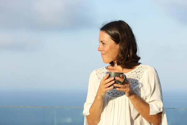一个严肃的成年女子在海滩的阳台上举着咖啡杯 望着别处 — 图库照片