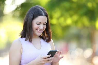 Parkta yürüyen mutlu bir kadın akıllı telefon içeriğini kontrol ediyor.