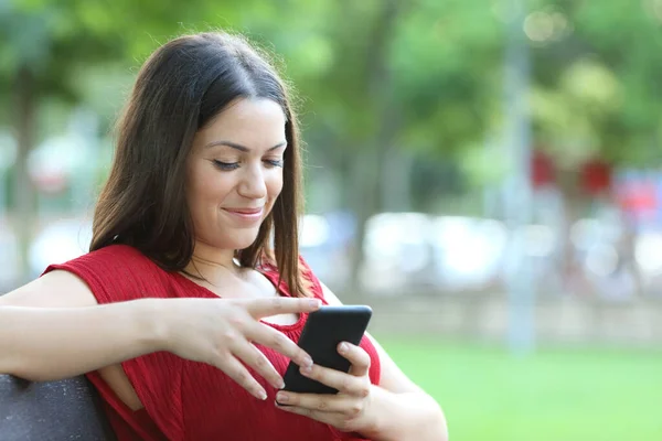 笑顔女性で赤チェックスマートフォン座っている上にベンチで公園 — ストック写真