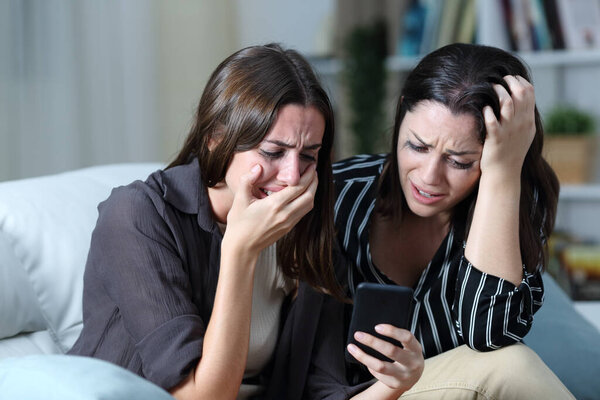 Sad friends or sisters crying checking smart phone on a couch in the living room at home 