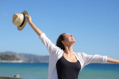 Güneşli bir günde kumsalda şapkasını tutan mutlu kadın kollarını çırpıyor.