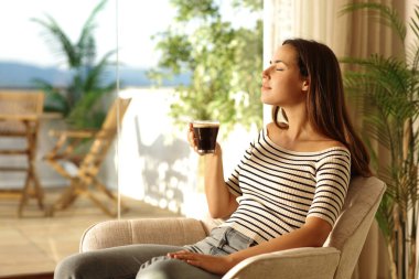 Kadın koltukta oturmuş, pencerenin yanında kahve içiyor.