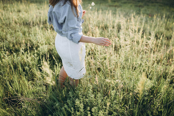 Girl in light linen clothes touching green grass in field at sunset in summer