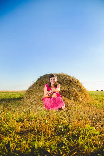 Girl in bright dress sitting in golden wheat field at a haystack on sunset and blue sky background. Rural scene, freedom and relaxation concept.