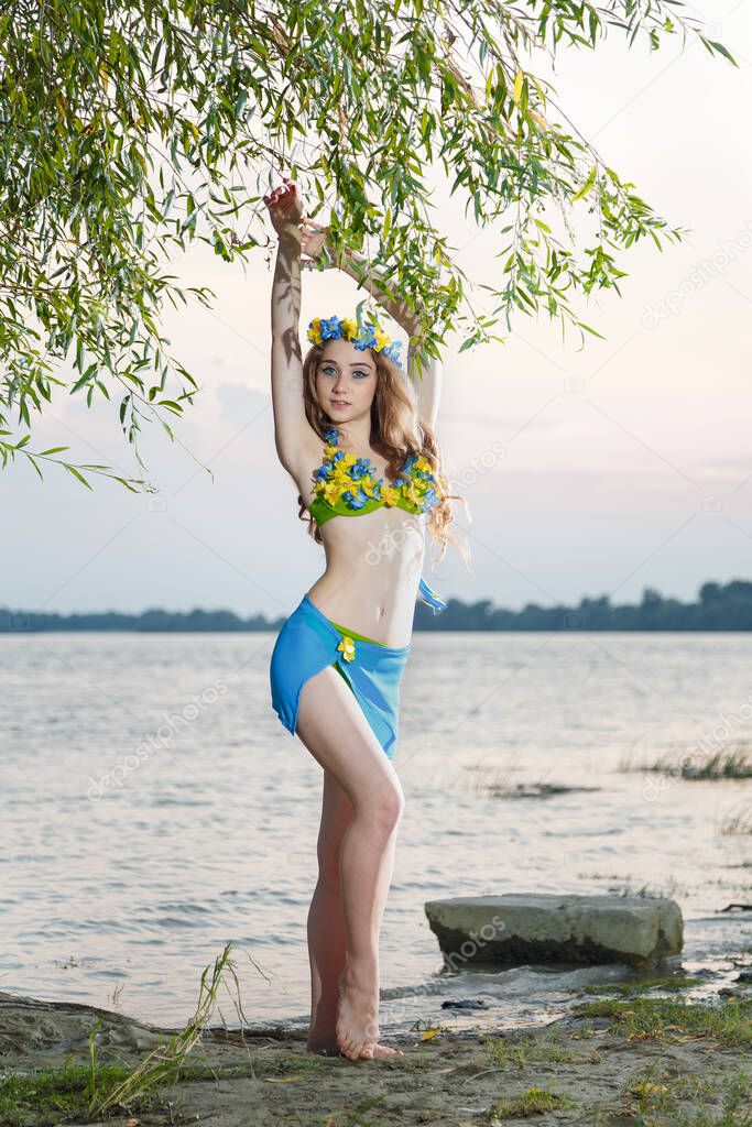 Ukrainian model in traditional flag colored swimsuit with flowers, wreath with ribbons, standing at river bank. Ethnic Ukrainian costume colors. 