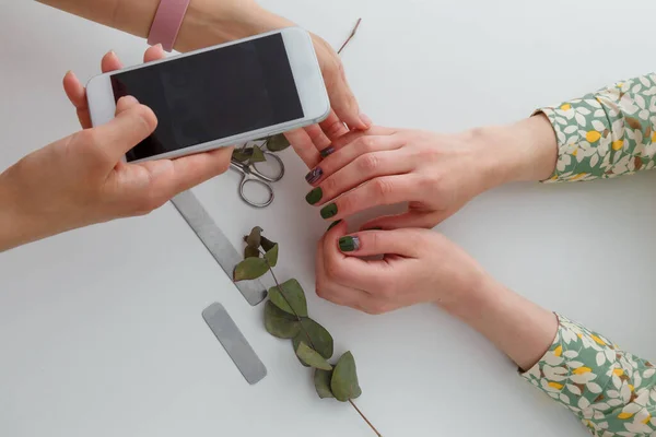 Nail artist taling photo of her work with mobile phone, only hands visible. Green fingernails design, nail files, manicure scissors and eucalyptus branch on white table. Beauty concept.