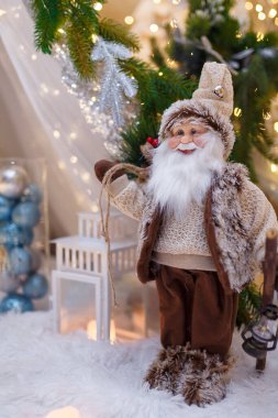 Kahverengi elbiseli Noel Baba oyuncağı beyaz fenerin önünde duruyor. Firavun ağacı dalları ve bulanık arka planda sıcak çelenk ışıkları ve bokeh ile Noel süslemeleri. Kış tatili konsepti. Dikey çekim.