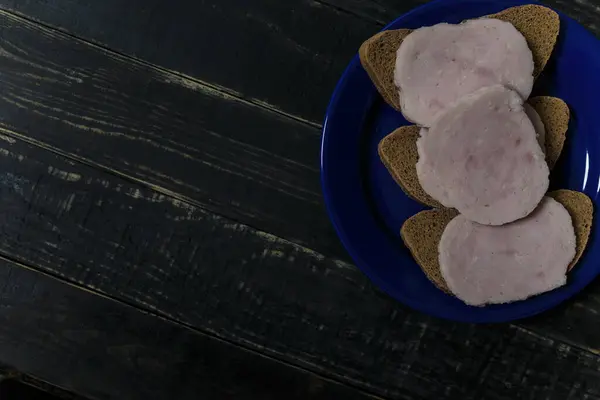 黒い木の背景に立って青いプレートの上に濃いパンとソーセージのスライスのサンドイッチ おいしい不健康な食べ物 食習慣の概念 テキストの場所 — ストック写真