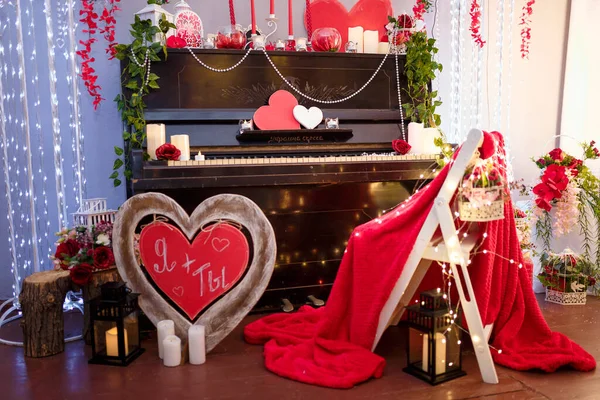 Valentine`s Day concept. Piano decorated with candles, red wooden hearts and flowers. Unfocused chair with red plaid and inscription Me + You on the foreground.