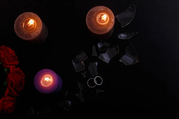 Love triangle and broken relationship concept. Wedding rings and three burning candles on black background. Flat lay, top view, place for text.