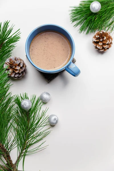 Blue cup of hot cacao or cappuccino with cinnamon standing on white table with pine branches and silver bulbs. Merry Christmas, Happy New Year and winter holidays concept. Copy space. Vertical shot.