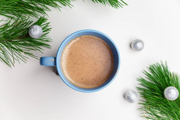 Blue cup of hot cacao or cappuccino with cinnamon standing on white table with pine branches and silver bulbs. Merry Christmas, Happy New Year and winter holidays concept. Copy space.