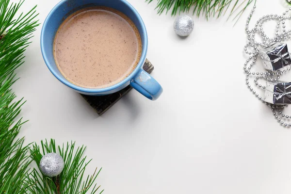 Blue cup of hot cacao or cappuccino with cinnamon standing on white table with pine branches and silver bulbs. Merry Christmas, Happy New Year and winter holidays concept. Copy space.