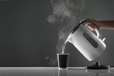 Electric white kettle pouring water in cup on table on grey background clipart