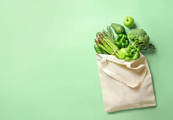 Eco bag full of different green fresh vegetables and herbs on light green background. Top view.