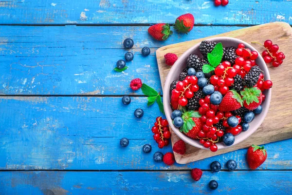 Ceramic bowl with assorted wild fresh berries on wooden cutting board on blue wooden background. Top view. Copy space.