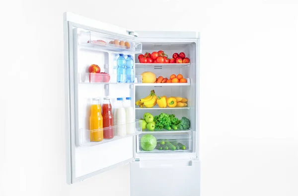Open fridge full of vegetables, fruits and drinks on white wall background.