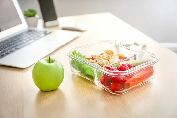 Healthy business lunch in office workplace. Vegetable and chicken salad in lunch box and fresh green apple on wooden desk near laptop.