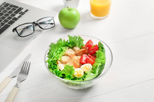 Healthy business lunch in office workplace. Vegetable and chicken salad in bowl, fresh green apple, glass of orange juice on white wooden desk near laptop and glasses.