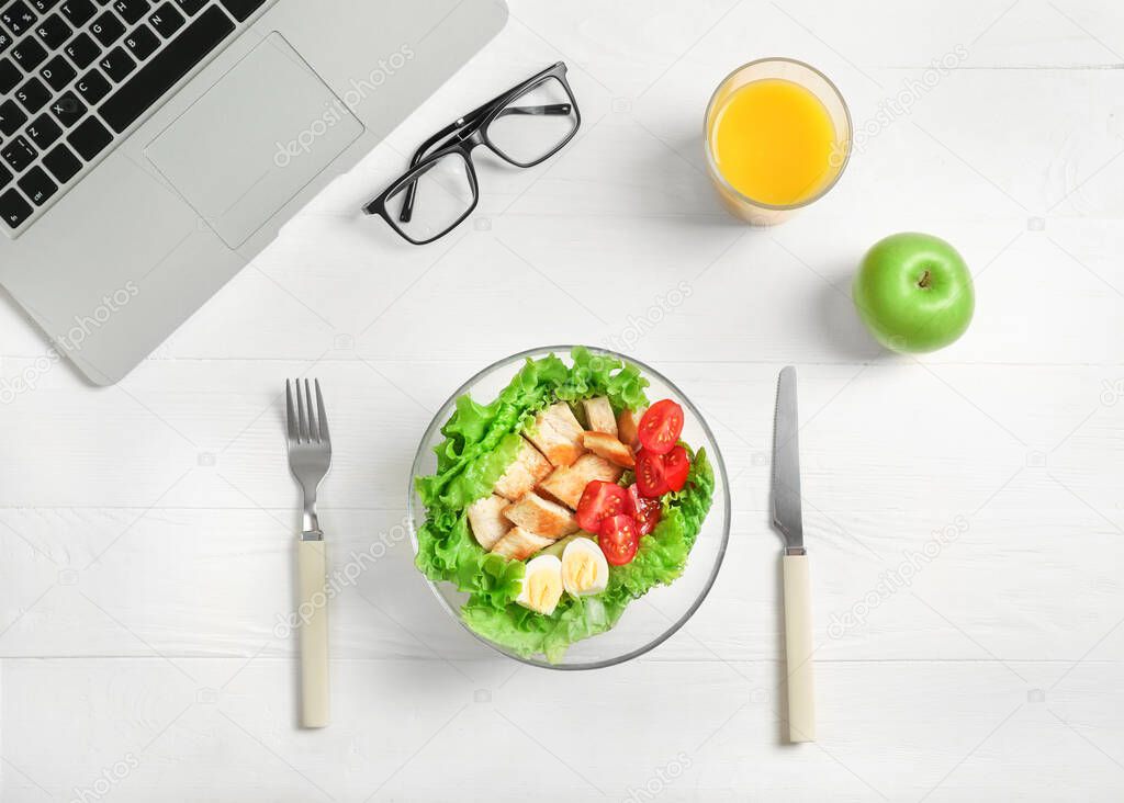 Healthy business lunch in office workplace. Top view of vegetable and chicken salad in bowl, fresh green apple, glass of orange juice on white wooden desk near laptop and glasses.