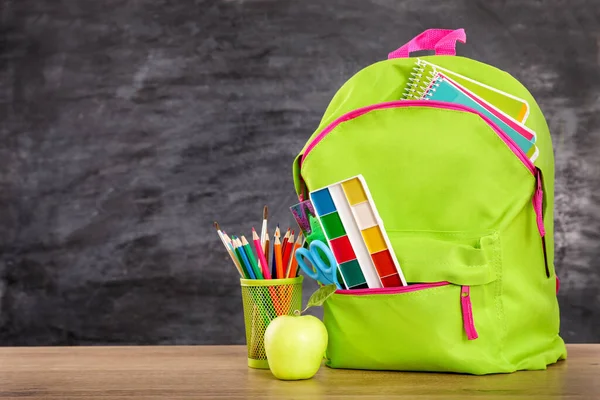 Green school bag full with school supplies on wooden desk over black chalkboard background. Copy space. Back to school concept.