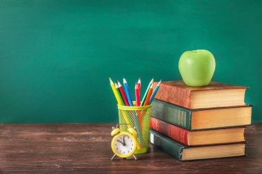 Yığınla kitap, elma, saat ve kalem standındaki renkli kalemler tahta masanın üzerinde yeşil karatahta arka planda. Boşluğu kopyala.