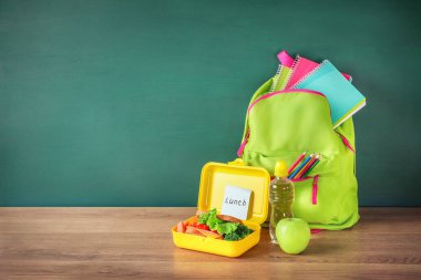 Yeşil okul çantası ve sağlıklı beslenme çantası ahşap masa üzerinde yeşil karatahta arka planda. Boşluğu kopyala.