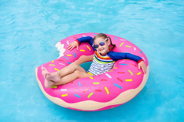 Child in swimming pool on funny inflatable donut float ring. Little girl learning to swim in outdoor pool of tropical resort. Water toys for kids. Healthy sport activity for children. Sun protection. 