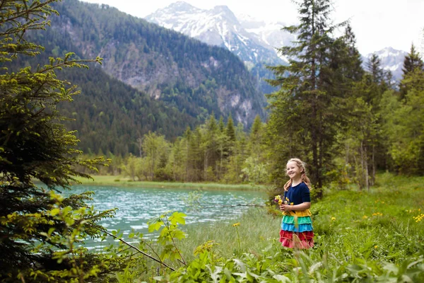 Child hiking in the Alps mountains looking at beautiful lake. Kid in alpine flower field at snow covered mountain in Austria. Spring family vacation. Little girl on scenic hike trail. Outdoor fun.