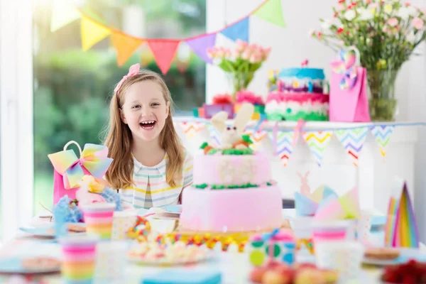 Kids birthday party with colorful rainbow pastel decoration and bunny layer cake. Little girl with sweets, candy and fruit. Balloons and banner at festive decorated table for child or baby birthday.