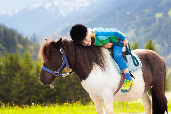 Kids riding pony in the Alps mountains. Family spring vacation on horse ranch in Austria, Tirol. Children ride horses. Kid taking care of animal. Child and pet. Little boy in saddle on pony.