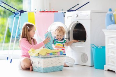 Children in laundry room with washing machine or tumble dryer. Kids help with family chores. Modern household devices and washing detergent in white sunny home. Clean washed clothes on drying rack. 