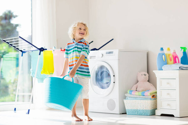 Child in laundry room with washing machine or tumble dryer. Kid helping with family chores. Modern household devices and washing detergent in white sunny home. Clean washed clothes on drying rack. 