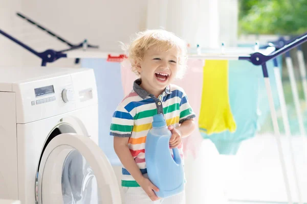 Child in laundry room with washing machine or tumble dryer. Kid helping with family chores. Modern household devices and washing detergent in white sunny home. Clean washed clothes on drying rack.