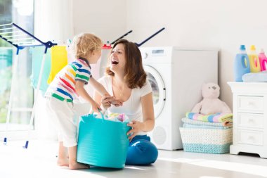 Mother and kids in laundry room with washing machine or tumble dryer. Family chores. Modern household devices and washing detergent in white sunny home. Clean washed clothes on drying rack. 