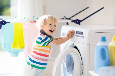 Çocuk Çamaşırhane, çamaşır makinesi ile Oda veya kurutma. Çocuk ile aile işleri yardımcı. Modern ev aygıtları ve çamaşır deterjan beyaz güneşli evinde. Raf kurutma üzerinde temiz yıkanmış kıyafetler. 