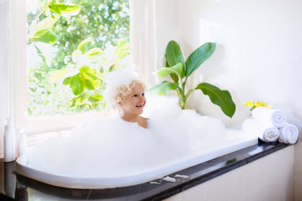 Little child taking bubble bath in beautiful bathroom with big garden view window. Kids hygiene. Shampoo, hair treatment and soap for children. Kid bathing in large tub. Baby boy with foam in hair.