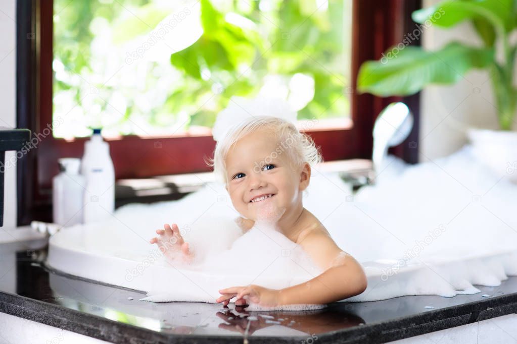 Little child taking bubble bath in beautiful bathroom with big garden view window. Kids hygiene. Shampoo, hair treatment and soap for children. Kid bathing in large tub. Baby boy with foam in hair.