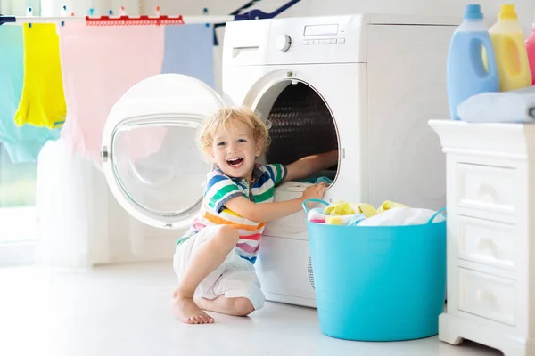 Child in laundry room with washing machine or tumble dryer. Kid helping with family chores. Modern household devices and washing detergent in white sunny home. Clean washed clothes on drying rack.