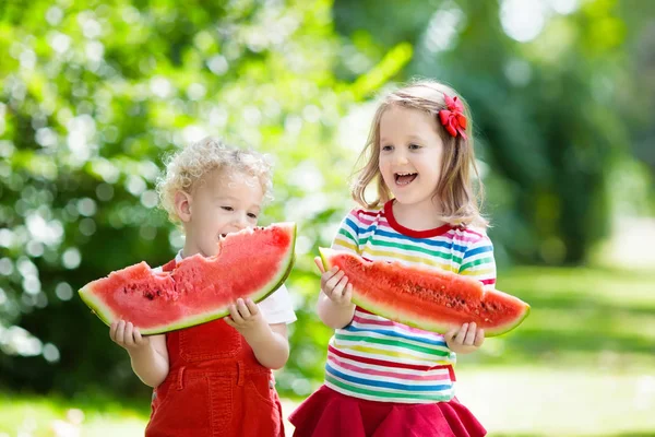 Child eating watermelon in the garden. Kids eat fruit outdoors. Healthy snack for children. Little girl and boy playing in the garden biting a slice of water melon.