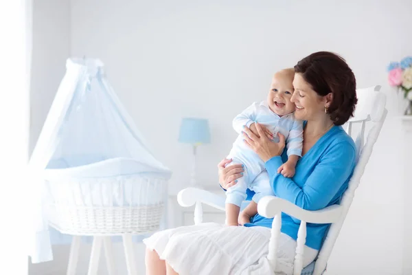 Young mother holding her newborn child. Mom nursing baby. Woman and new born boy in white bedroom with rocking chair and blue crib. Nursery interior. Mother playing with laughing kid. Family at home