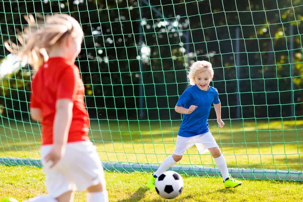 Kids play football on outdoor field. Children score a goal at soccer game. Girl and boy kicking ball. Running child in team jersey and cleats. School football club. Sports training for young player.