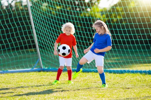 Kids play football on outdoor field. Children score a goal at soccer game. Girl and boy kicking ball. Running child in team jersey and cleats. School football club. Sports training for young player.